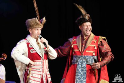 Dwóch artystów w barwnych, historycznych strojach szlacheckich z piórami w czapkach występuje na scenie. Jeden z nich mówi do mikrofonu, drugi śmieje się szeroko, trzymając mikrofon w dłoni.